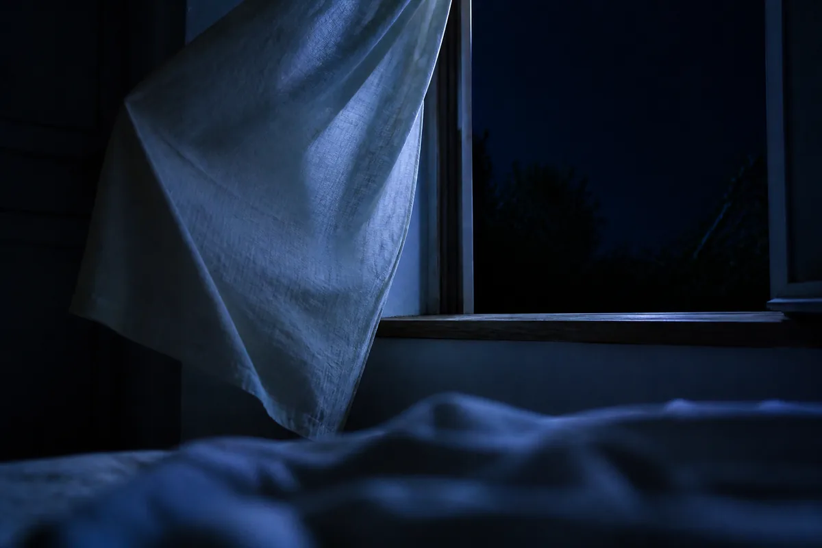 Fenêtre ouverte la nuit sur une chambre sombre, voilage de lin soulevé par un courant d&rsquo;air
