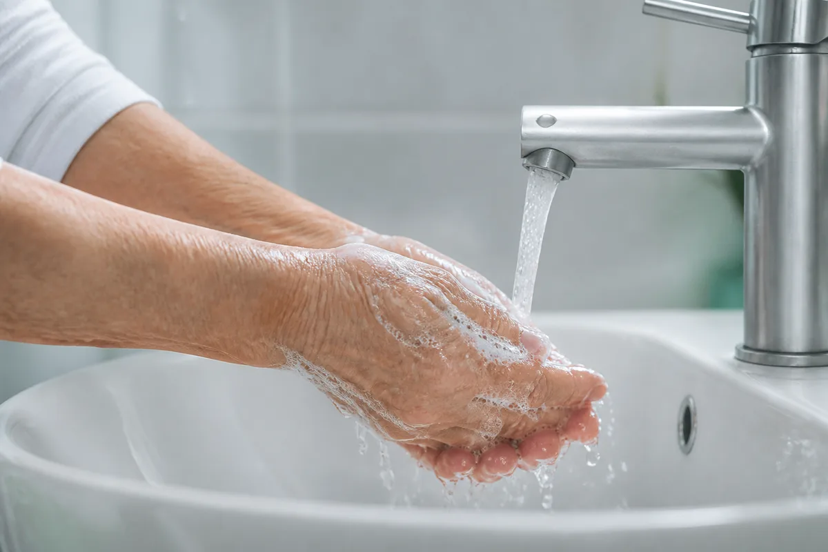 Mains mûres couvertes de mousse rincées sous un filet d’eau tiède au-dessus d’une vasque en céramique blanche